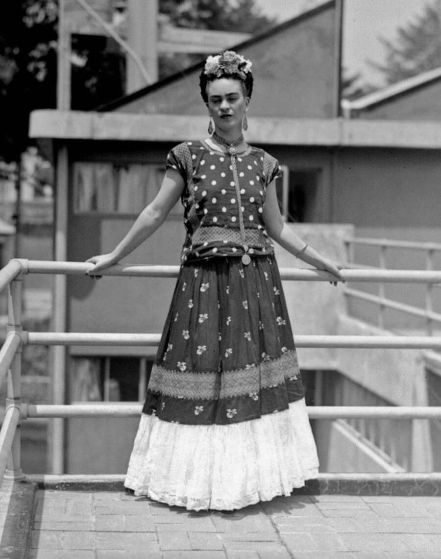 Frida Kahlo parada sostiene un barandal, vestida con falda larga, una bluda de lunares, un collar y flores en la cabeza