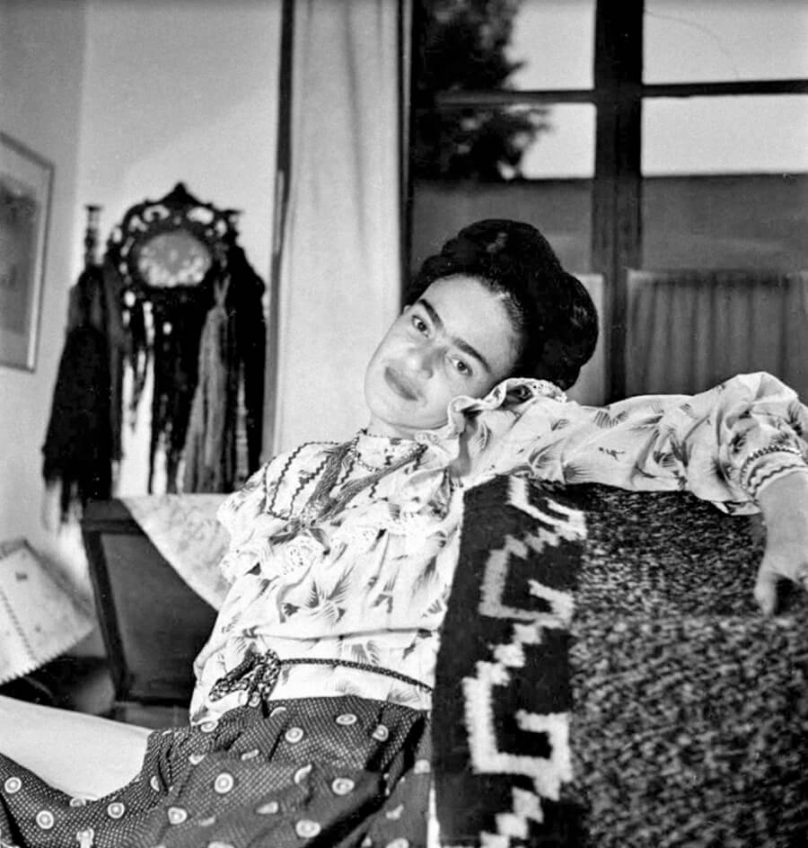 Frida Kahlo sentada en un sofá con los brazos extendidos y recargada sobre su hombro izquierdo