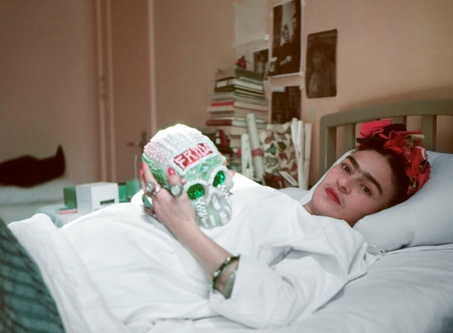 Frida Kahlo acostada en una cama sostiene con un cráneo de azúcar que lleva su nombre en la frente
