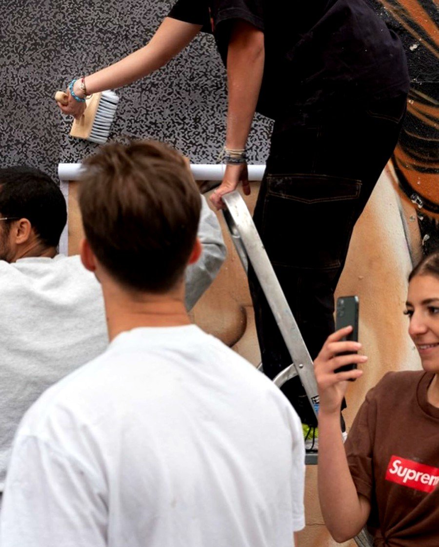 person painting JR's mural in Paris while a woman takes a picture with her cellphone